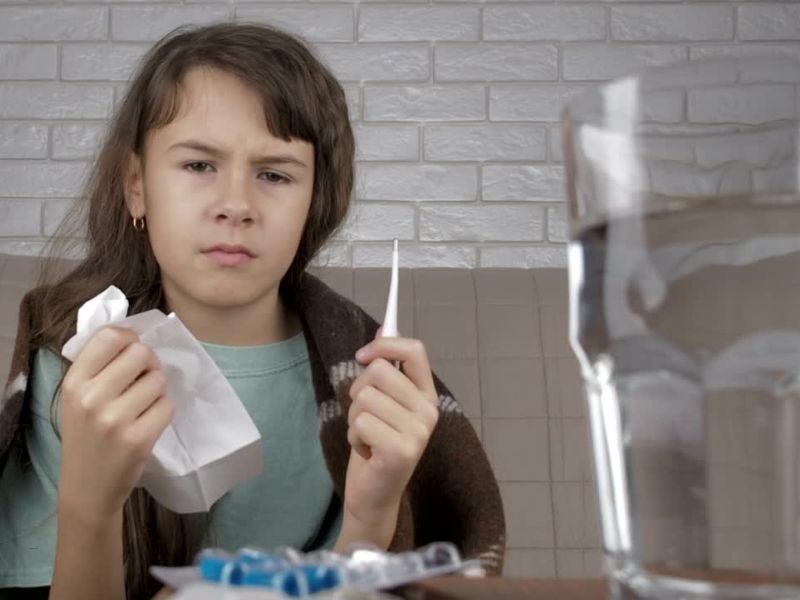 devojčica koja pati od prehlade drži toplomer u urci i maramice, na stolu voda i lekovi