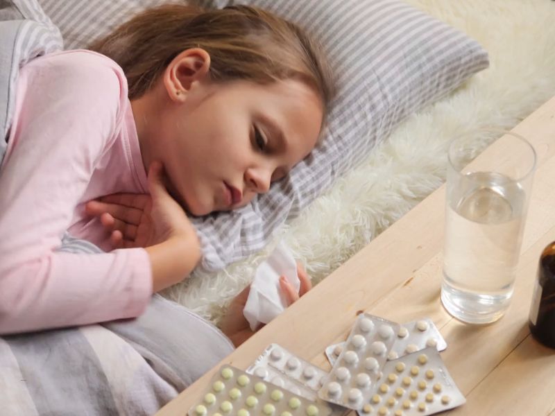 devojčica leži bolesna u krevetu, na stolčiću pored nje lekovi, voda i sirup