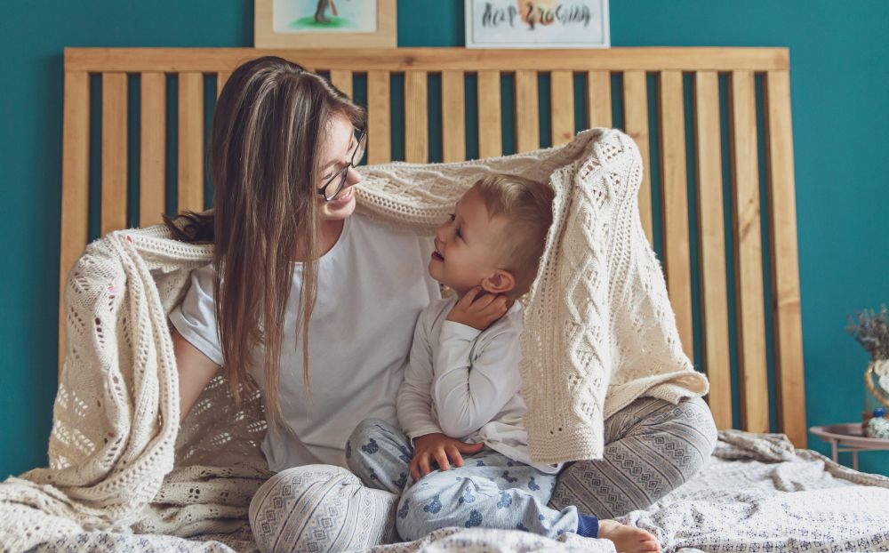 Majka i malo dete sede na krevetu, razgovaraju i osmehuju se tokom mirnog jutarnjeg buđenja